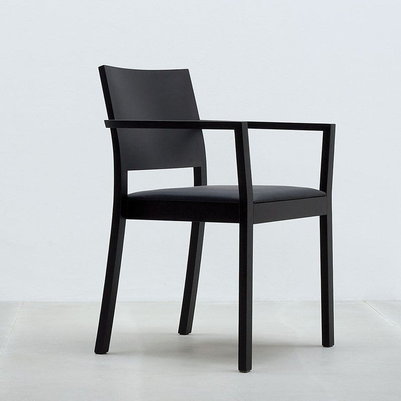 The ST4N Armchair by Hussl is a sleek, minimalist black chair crafted from solid wood, featuring clean lines and a robust backrest. It includes armrests and a flat seat, blending contemporary style with ergonomic comfort against a light gray background.