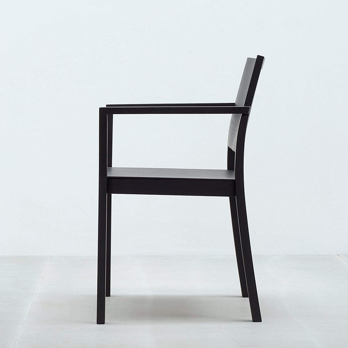 The ST4N Armchair by Hussl, featuring a minimalist black wooden design with ergonomic backrest and armrests, sits on a light gray floor against a pale gray wall.