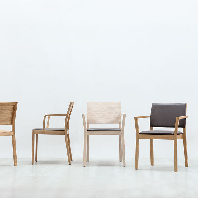 Four modern Hussl ST4N chairs with armrests are lined up against a plain white backdrop. Each is crafted from solid wood, featuring an ergonomic design with unique styles and colors, from light wood with gray seats to dark wood with black seats.