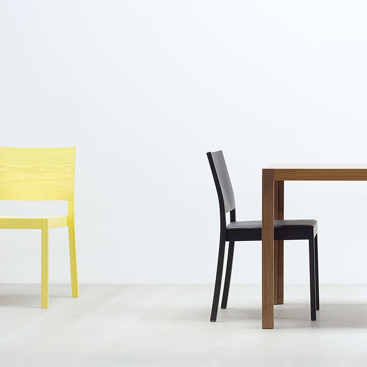 A minimalistic room showcases a solid wood table with Hussls ergonomic black ST4N Chair to the right and a separate yellow chair on the left against a white wall, complemented by a light gray floor that enhances the clean, modern aesthetic.
