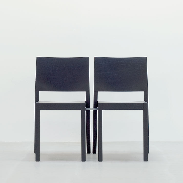 Two black ST4N Chairs by Hussl, made from solid wood with minimalist straight backs and legs, create a symmetrical and modern look against a plain light-colored wall.
