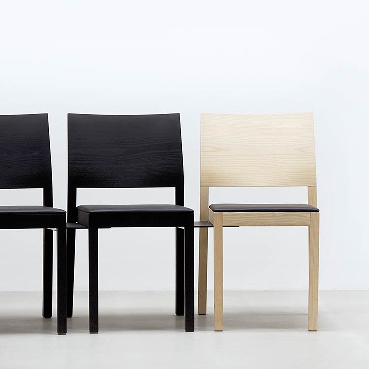 Four ST4N chairs by Hussl line up against a white wall: three black and one in solid wood with a light finish. All feature simple, modern designs with flat seats and slightly angled backs for an ergonomic experience.