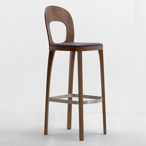 The ST6N-1 Barstool by Hussl is a modern wooden seat inspired by Austrian coffee house designs, featuring a cut-out backrest and dark cushion. It stands on four slim legs with a rectangular metal footrest against a light gray background.