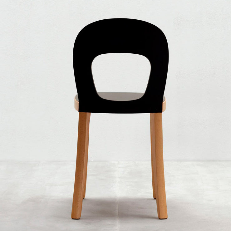 The ST6N-1 chair by Hussl features a sleek black backrest with an oval cutout design, inspired by Austrian coffee house style. It has a stable flat brown seat supported by four light wooden legs, set against a plain white wall and light-colored floor.