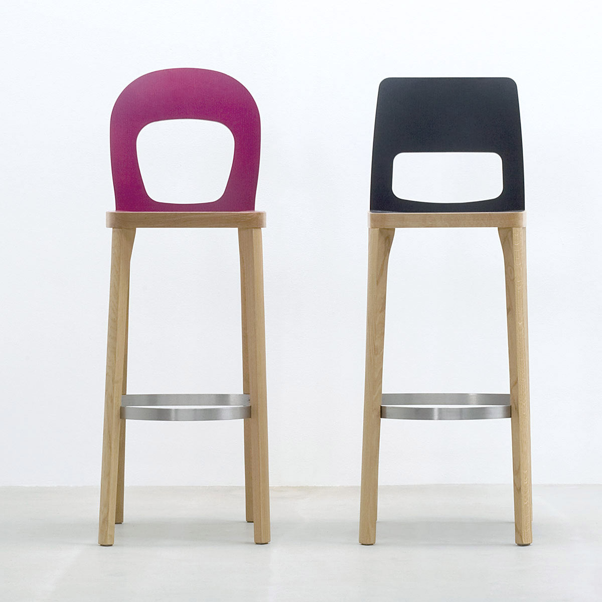 Two ST6N-2 Barstools by Hussl, inspired by Austrian coffee house designs, stand on a light floor against a white wall. Featuring oak legs and metal footrests, one stool has a pink backrest and the other black, both with cut-out designs.