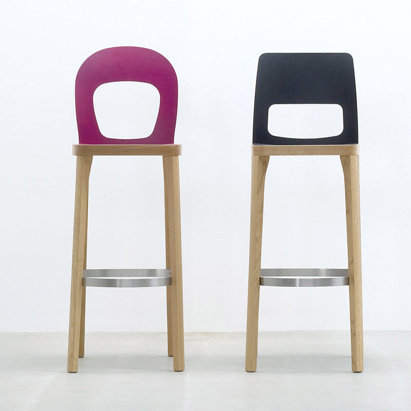 Two ST6N-2 Barstools by Hussl, inspired by Austrian coffee house designs, stand on a light floor against a white wall. Featuring oak legs and metal footrests, one stool has a pink backrest and the other black, both with cut-out designs.