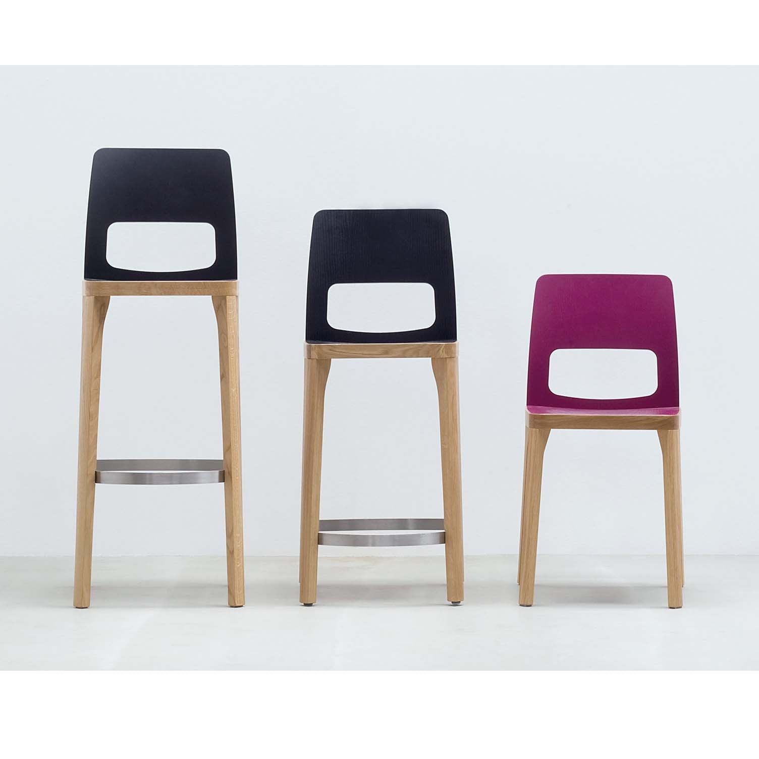 Against a plain white wall, two tall black and dark wood Hussl ST6N-2 Barstools feature metal footrests and cut-out backrest designs akin to Austrian café styles. The third is a shorter magenta chair with similar design elements, all boasting wooden legs.