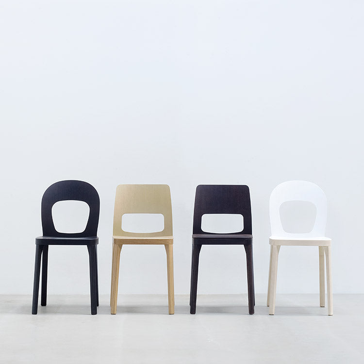 Four modern, minimalist ST6N-2 chairs by Hussl in black, beige, dark brown, and white are lined up on a light gray floor against a plain white wall. Each features an elegant backrest design with a round cutout, reminiscent of Austrian coffee house furnishings.