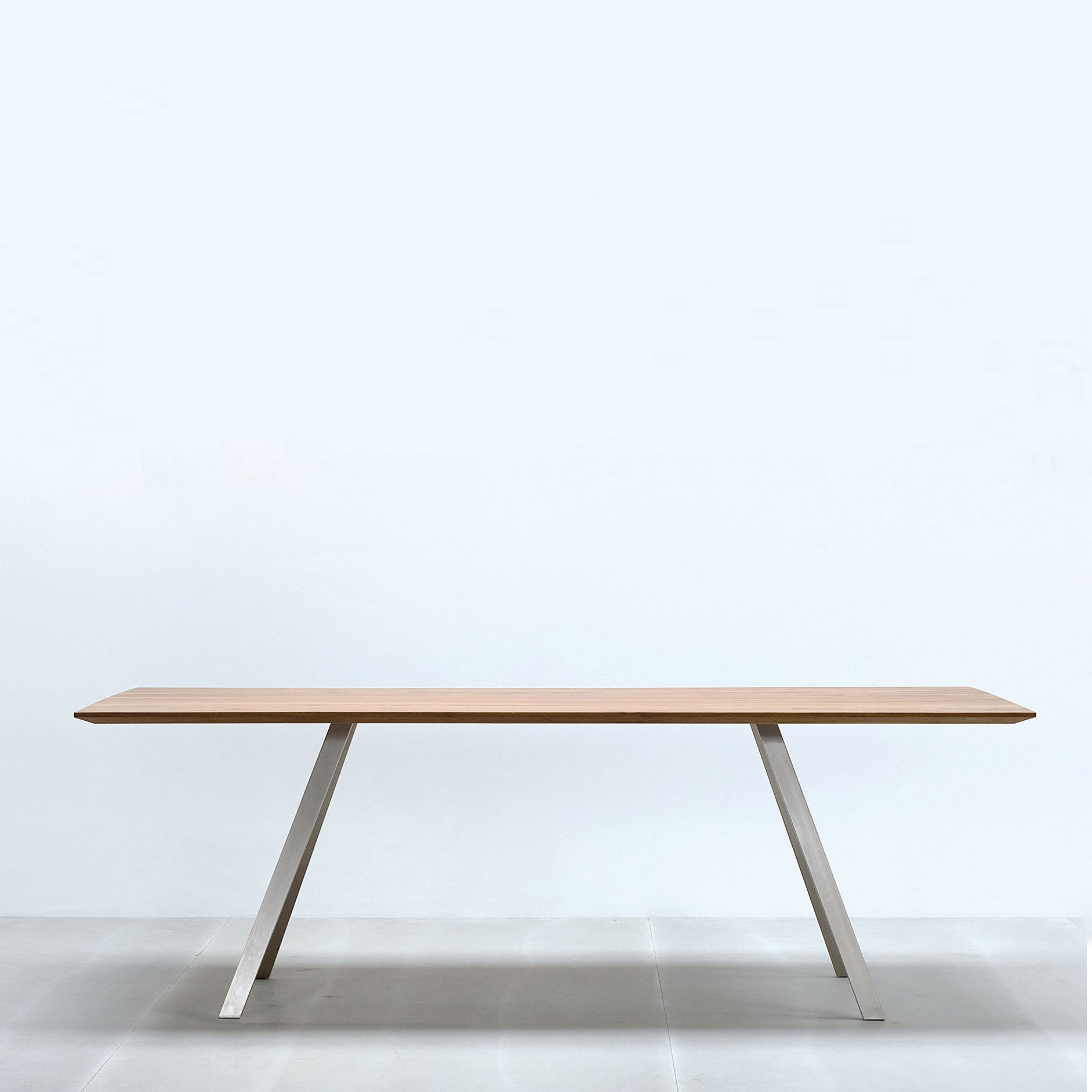 The Solid T Rectangular by Hussl is a sleek high table with a solid wood tabletop in light wooden tones and stylish angled metal legs, set against a minimalist light blue background.