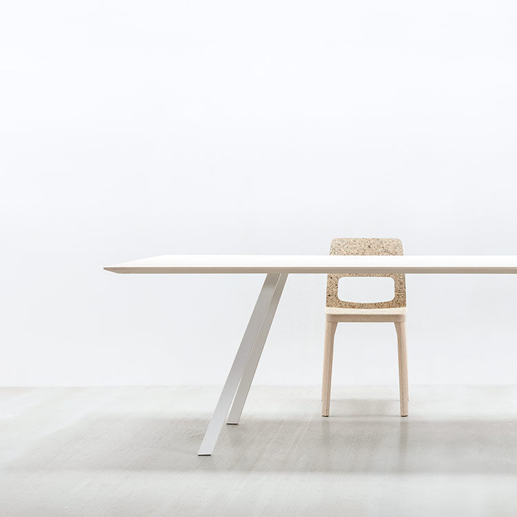 A minimalist room showcases the Hussl Solid T Rectangular table, featuring a light wooden top with angled metal legs, paired with a matching chair with a cutout backrest. The tables solid wood blends seamlessly with the smooth, light-colored floors and walls, highlighting simplicity and clean lines.