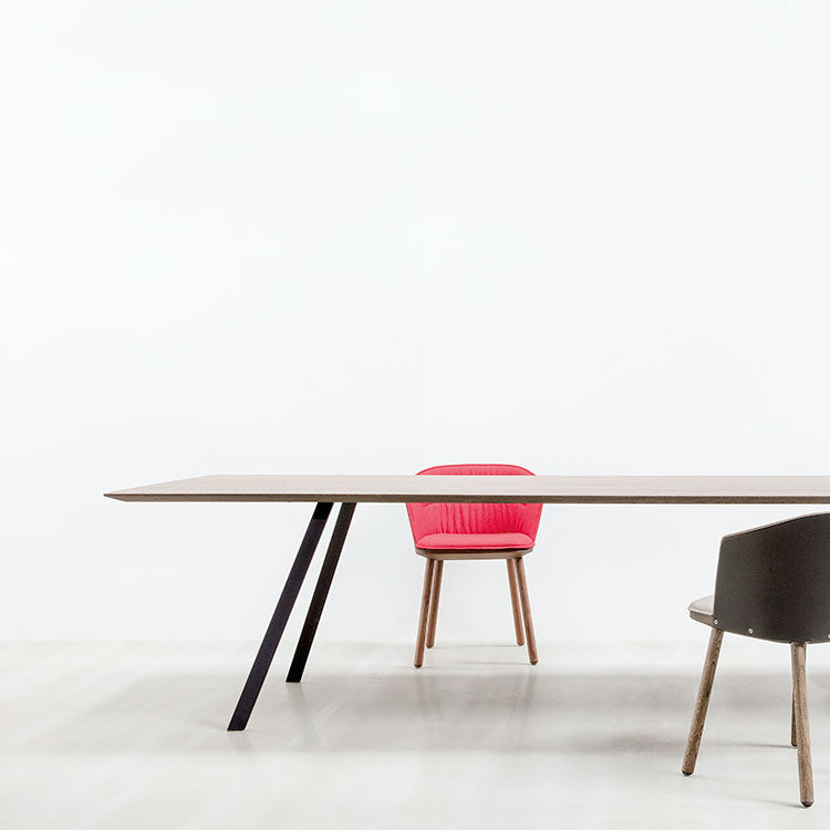 A minimalist scene features a Hussl Solid T Rectangular light wooden table with black angled legs. A red cushioned chair is centered behind, partly visible. To the right, a gray chair with wooden legs stands in profile against the plain white background.