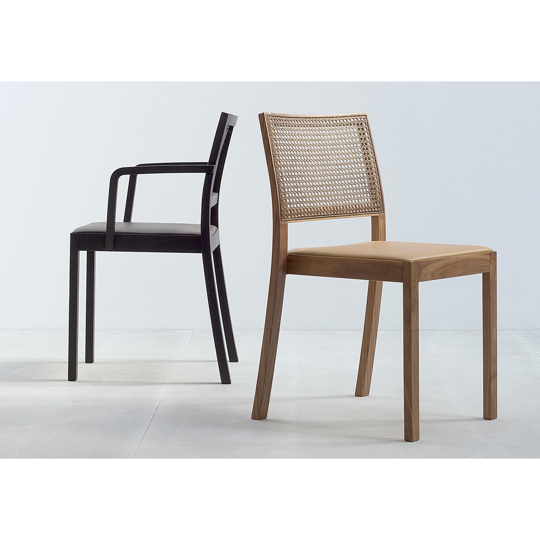 Two modern chairs on a light floor. The left features the ST4N Gritsch by Hussl, black with armrests and sleek coffeehouse design. To its right, another chair sports a wooden frame with a Viennese weave backrest and no armrests, both facing away from each other.