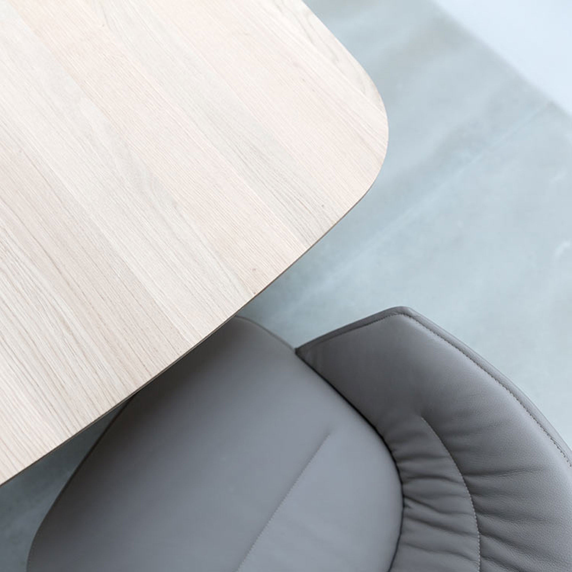 Top view of a light wooden table with rounded edges paired with the Hussl Lino C armchair, featuring an ergonomic design. The chair is partially beneath the table on a light gray floor, enhancing a minimalist and modern aesthetic with natural materials.
