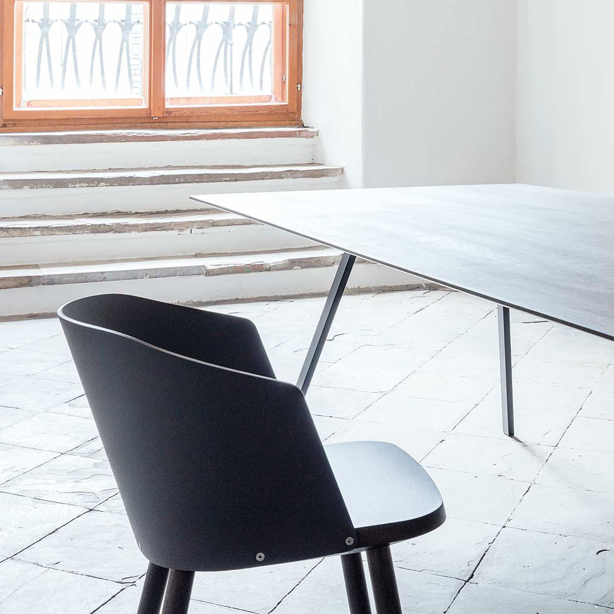In a modern minimalist room, a Hussl Lino C armchair sits next to a sleek dark rectangular table on white tile flooring. Sunlight filters through a window with decorative bars, casting soft shadows on the floor and stairs, accentuating the ergonomic design.