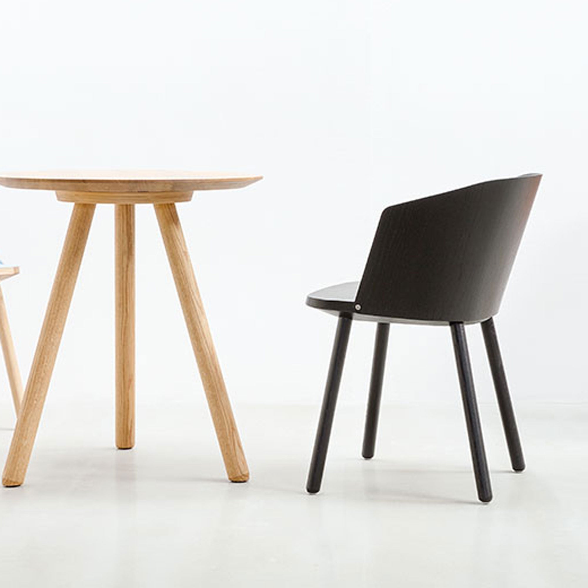 A minimalist scene showcases a wooden round table with three legs on the left and a Hussl Lino C armchair, featuring an ergonomic design and four straight legs, on the right. Both pieces are made from natural materials and set against a plain white background.