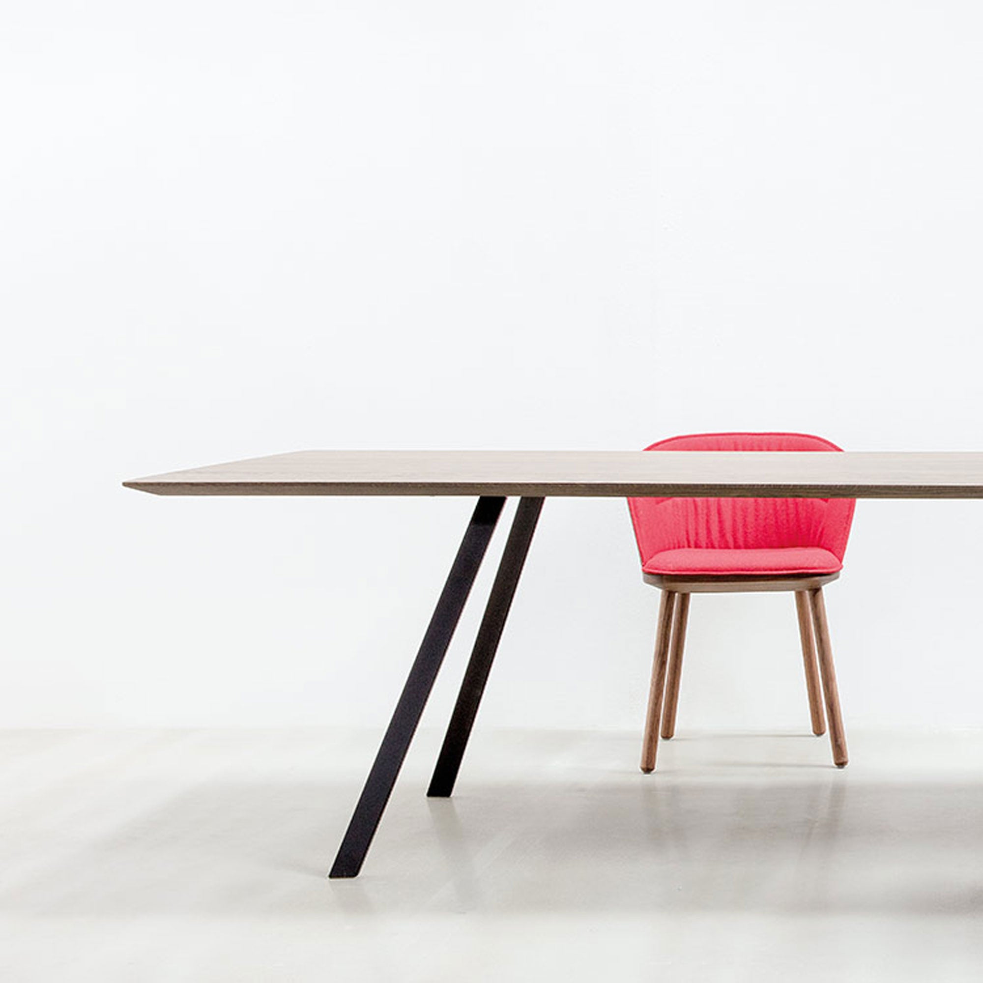 The setup showcases a modern table with angled black legs and a pink cushioned Lino C armchair by Hussl, featuring wooden legs partially tucked underneath. Crafted from natural materials, it stands out against a white background, emphasizing ergonomic design and minimalist elegance.