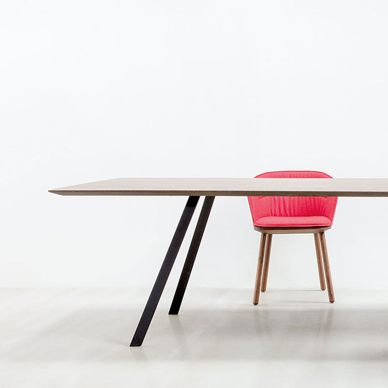 The setup showcases a modern table with angled black legs and a pink cushioned Lino C armchair by Hussl, featuring wooden legs partially tucked underneath. Crafted from natural materials, it stands out against a white background, emphasizing ergonomic design and minimalist elegance.