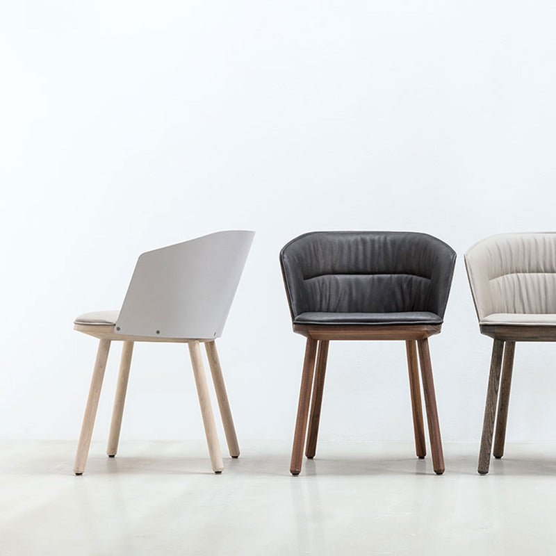 Three modern chairs are placed on a light floor against a white wall. The first is the Lino C armchair by Hussl with a minimalist design; the second chair is black with an ergonomic cushioned seat; the third is light gray, featuring tufted cushioning and made from natural materials.