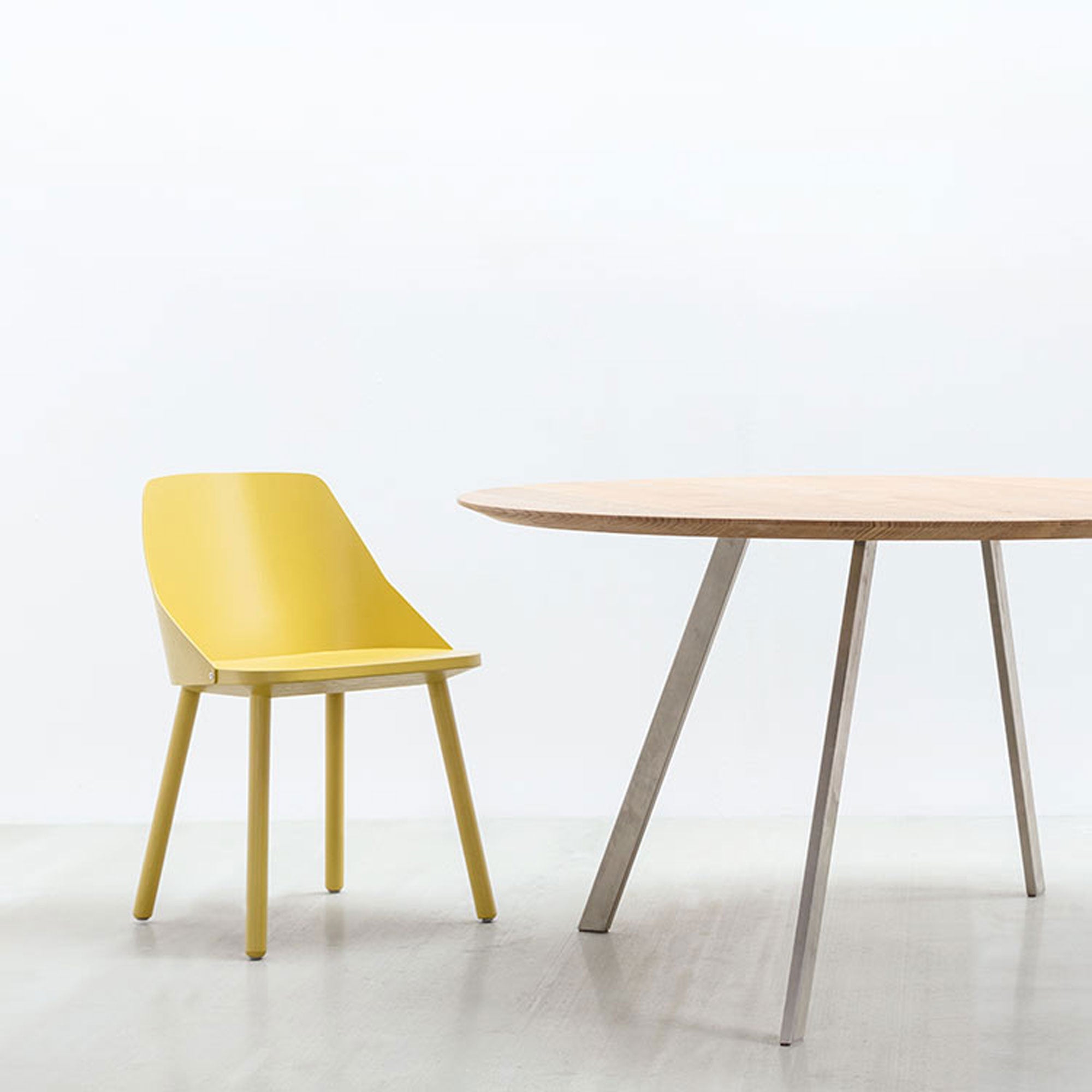 A minimalist dining scene features Hussls Lino C, a modern yellow shell chair with a curved backrest and elegant upholstery, beside a round wooden table with angled metal legs. The natural materials blend seamlessly against the plain white wall and light wood floor.