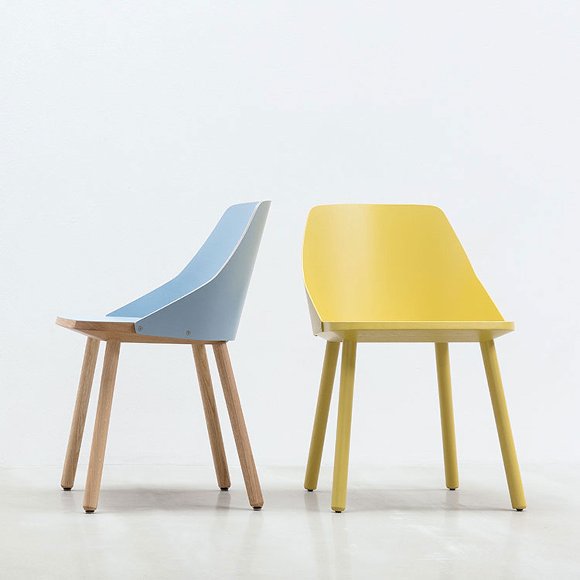 Two modern Hussl Lino C shell chairs on a light background: one has a light blue seat with wooden legs, showcasing natural materials; the other features a yellow seat with matching yellow legs. Both display minimalist design and refined upholstery.