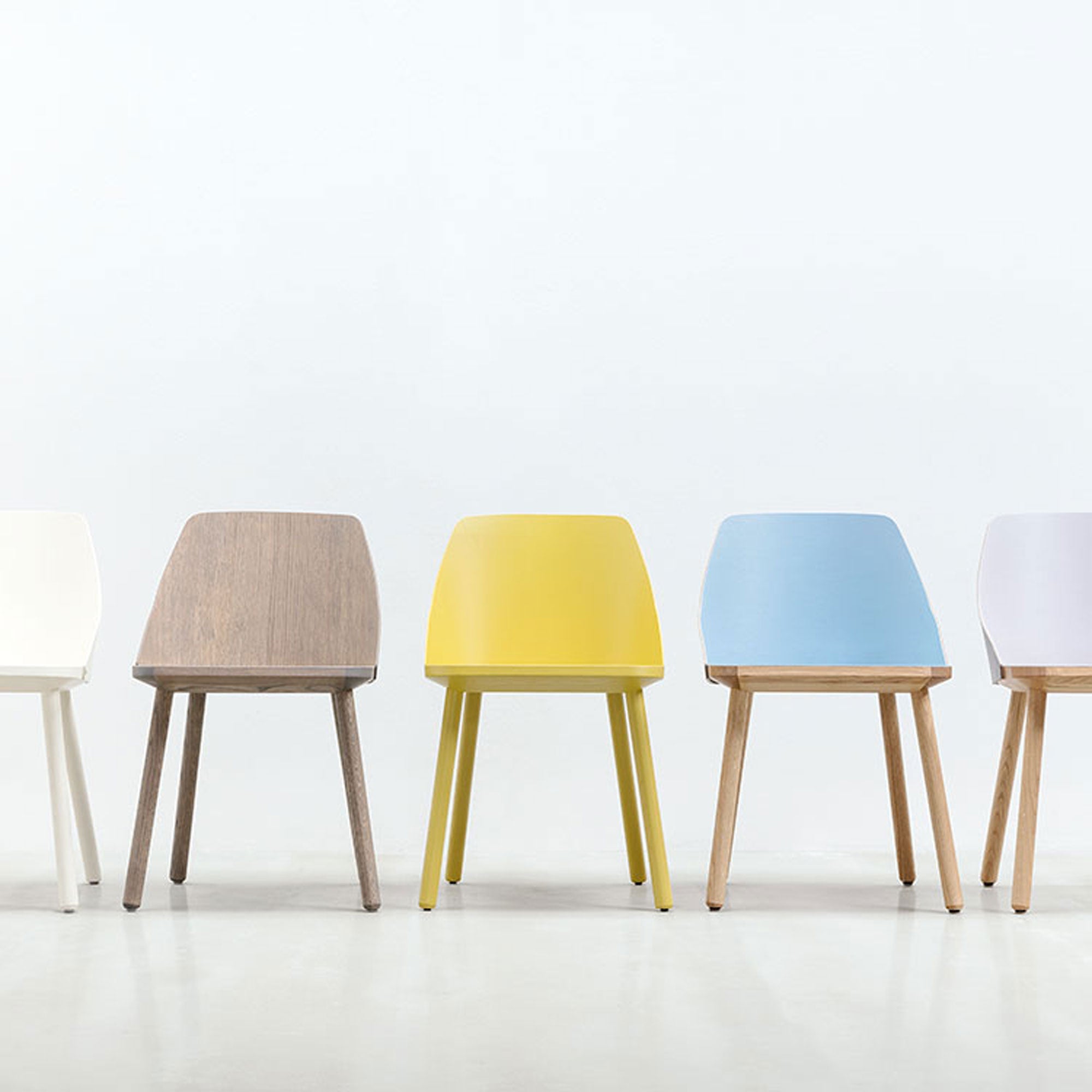 A row of Lino C chairs by Hussl in diverse colors—white, wood, yellow, blue, and another white—lines up against a plain backdrop. Their sleek design paired with wooden legs made from natural materials provides style and comfort.