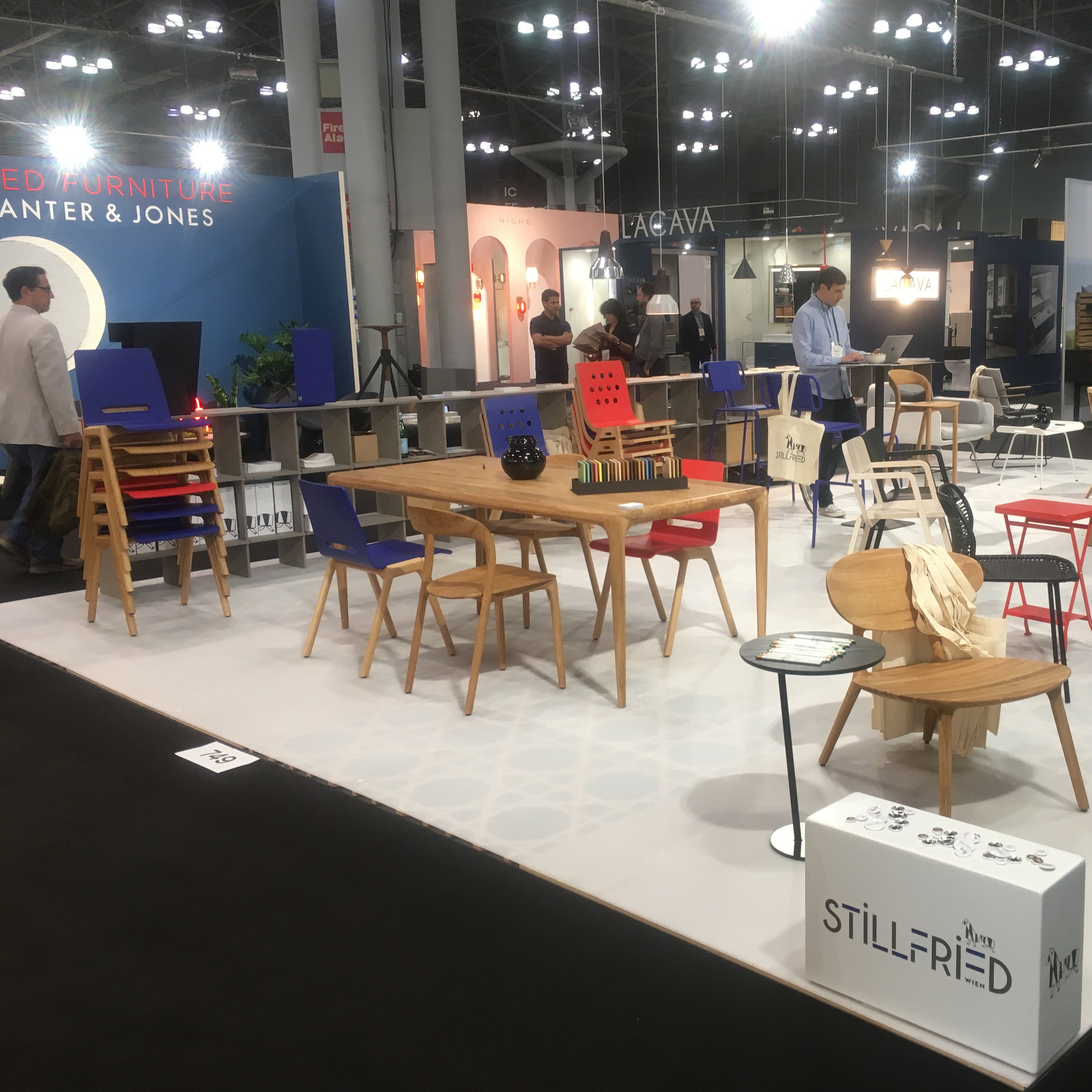 At a modern furniture exhibition booth, Hussls Forum 2S - Ultramarine Sale vibrant chairs are featured alongside wooden tables and decor on carpet. Visitors browse the displays under bright lights in a spacious indoor venue.