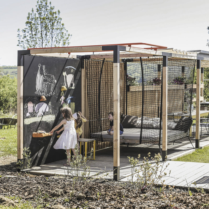 Children enjoy an outdoor Leva Home by Egoe, featuring chalkboard walls. A child in a white dress draws with chalk while another sits inside. The space is surrounded by greenery and wooden walkways, creating a serene and tranquil environment under the sunny sky.
