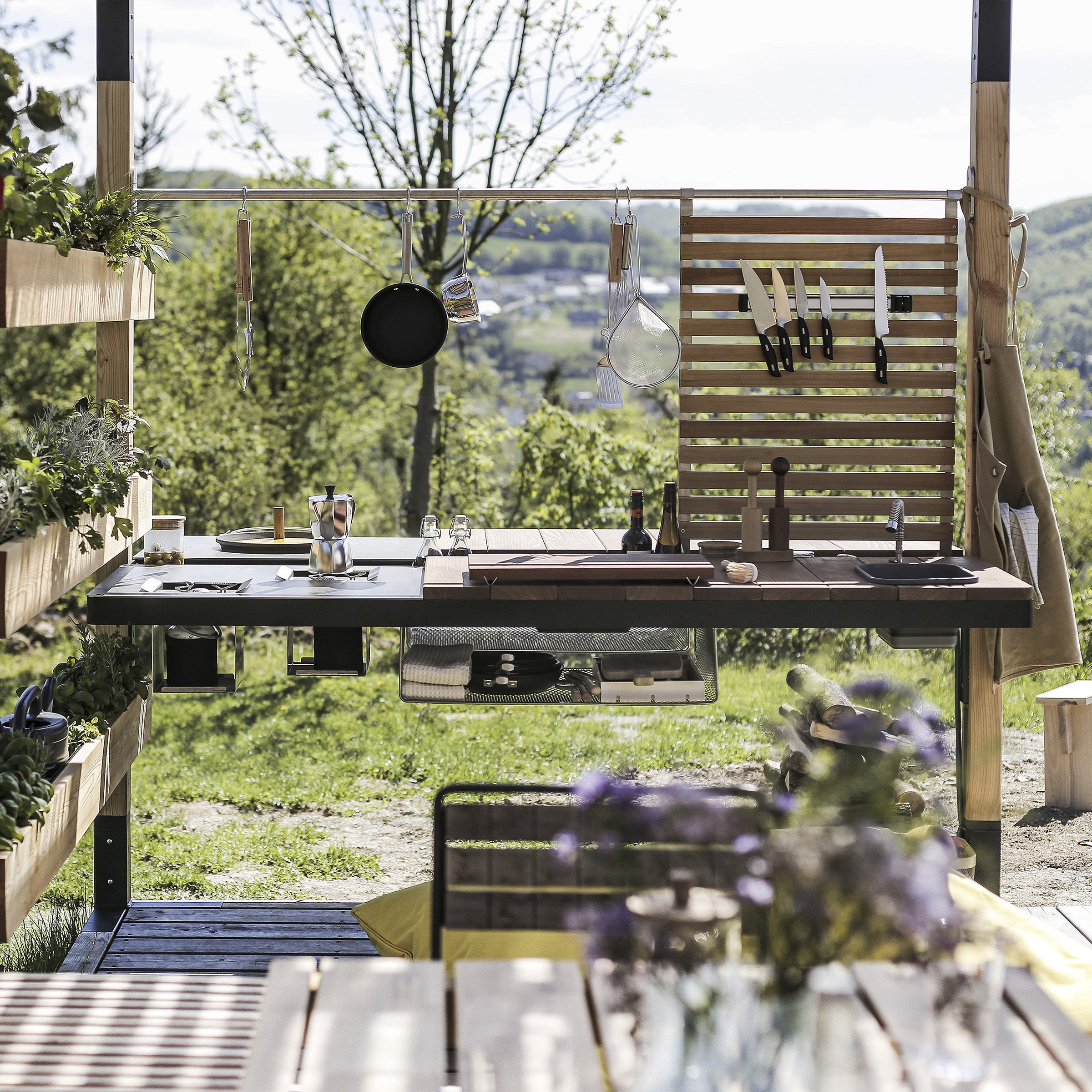 An outdoor kitchen with a wooden countertop, hanging pots and knives, surrounded by lush trees. Shelves are filled with plants and tools, while Egoes Leva Home modular system adds functionality. In the foreground, a table adorned with glasses and flowers invites relaxation.