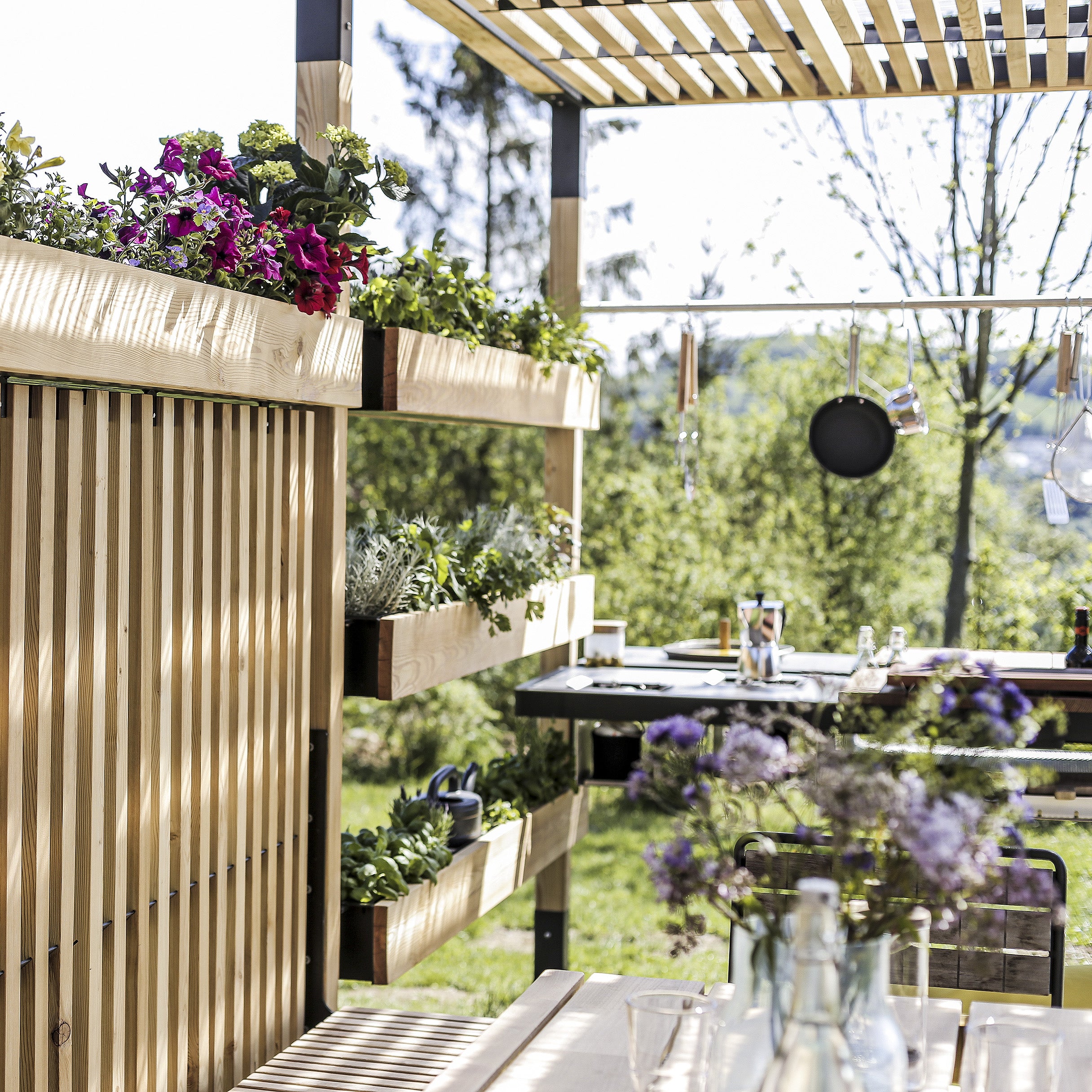 The outdoor kitchen with a wooden pergola and Egoes Leva Home system exudes tranquility. Lush planters, hanging utensils, and a wooden table adorned with flowers and glasses create a relaxing scene against green trees and serene mountains.