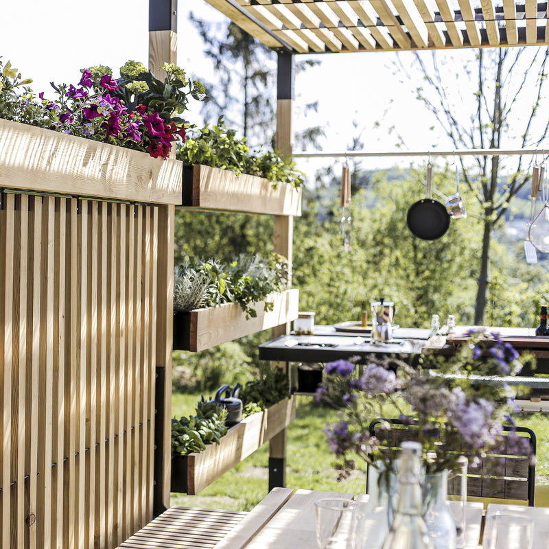 The outdoor kitchen with a wooden pergola and Egoes Leva Home system exudes tranquility. Lush planters, hanging utensils, and a wooden table adorned with flowers and glasses create a relaxing scene against green trees and serene mountains.
