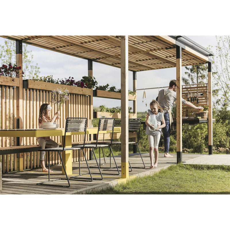 A family relishes an outdoor meal under a plant-covered wooden pergola with the Leva Home system by Egoe. A child sits at a yellow table, another strolls by, and someone grills. Surrounded by lush greenery in the sun, the scene exudes relaxation and tranquility.