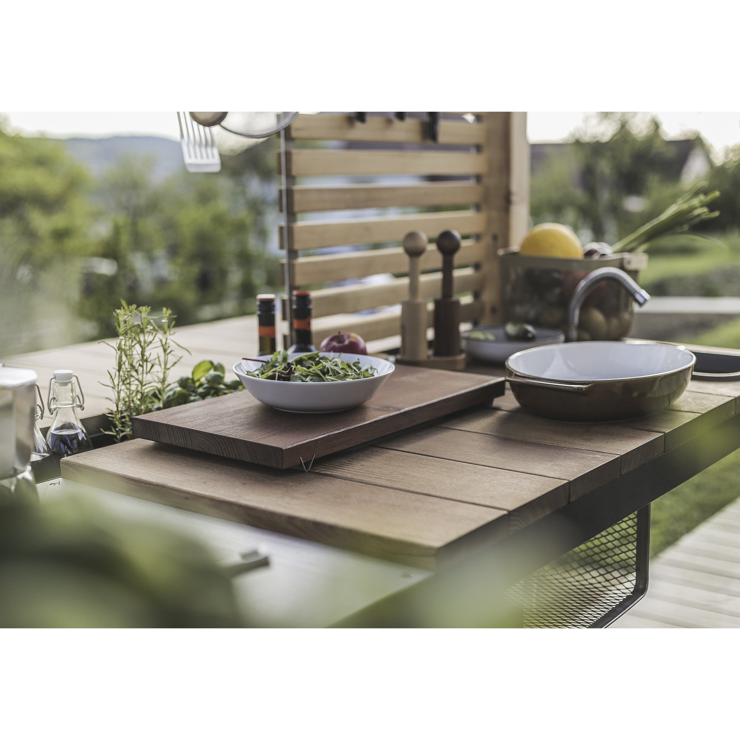 A Leva Home modular system by Egoe features prominently in an outdoor wooden kitchen counter, surrounded by bowls, fresh herbs, condiments, and cooking utensils. The tranquil scene includes blurred greenery and a wooden slatted rack as the serene backdrop.