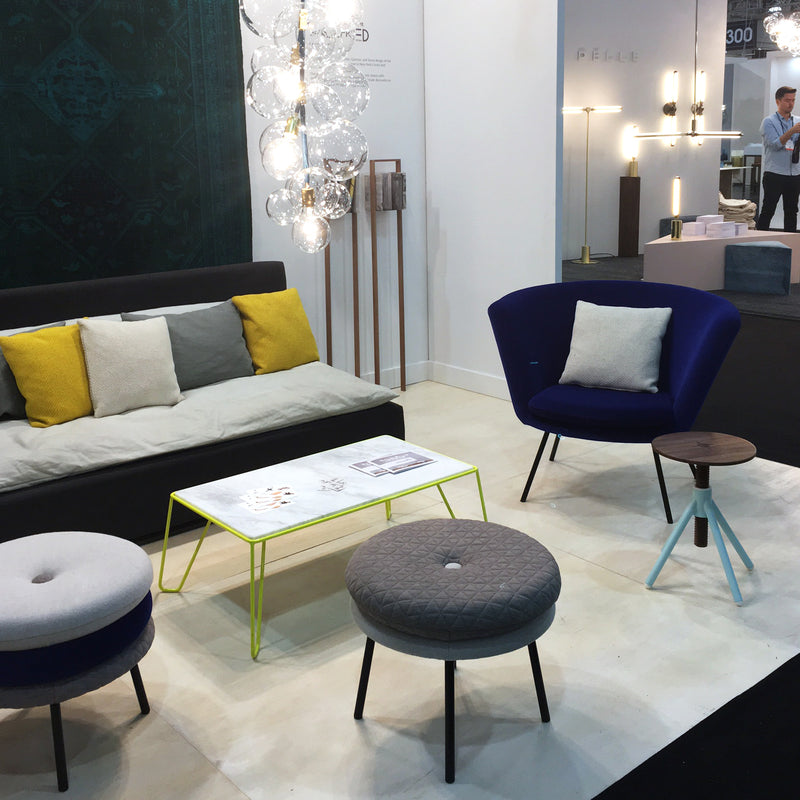 A modern living room features a dark sofa with yellow/gray pillows, a blue armchair, and the Little Tom Stool by Richard Lampert. A white coffee table with a yellow frame complements the space, while decorative hanging lights and floor lamps add ambiance.