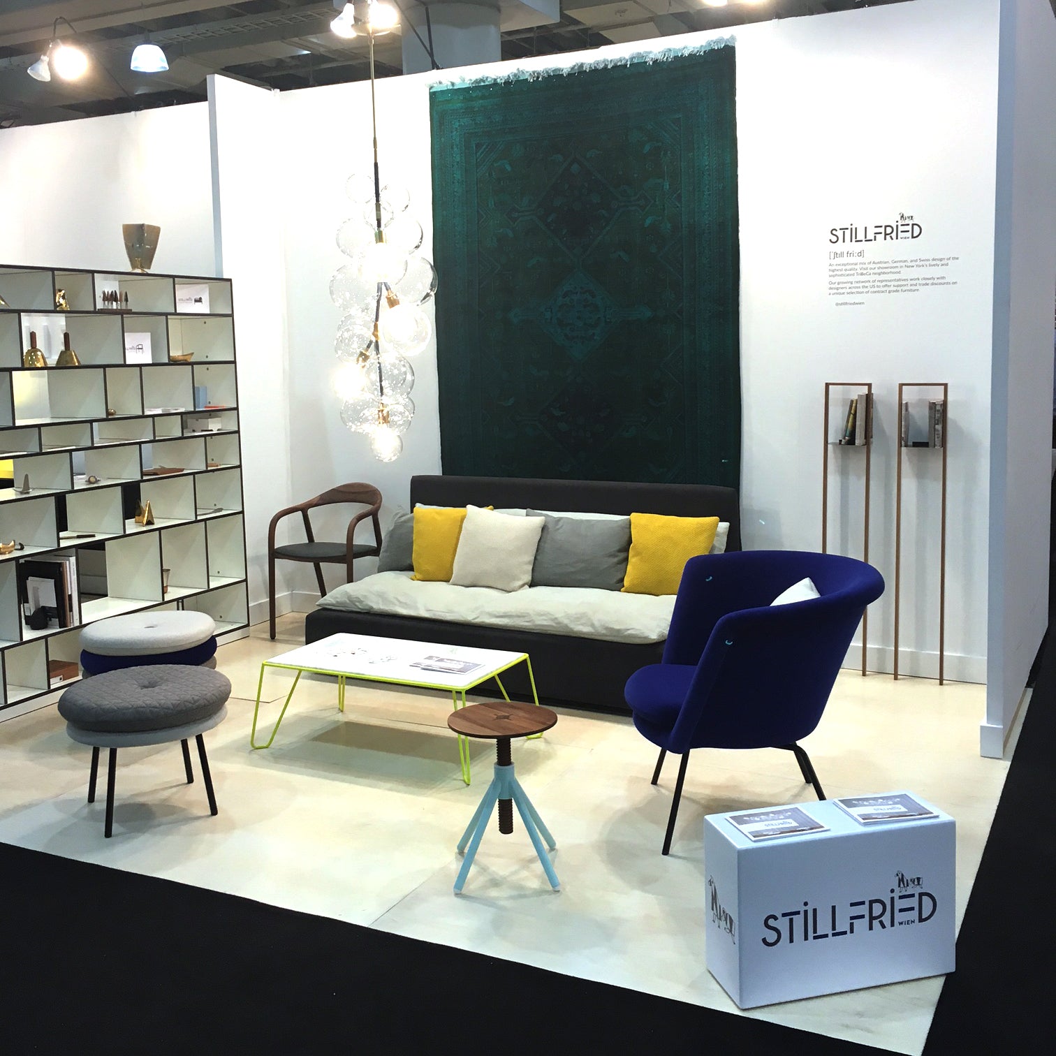 A showroom displays a gray couch with yellow and gray cushions, a blue chair, and various tables. A geometric shelf stands on the left, while Richard Lamperts Little Tom Stool by designer Alexander Seifried adds flair against a textured dark green wall hanging that contrasts with the white walls.