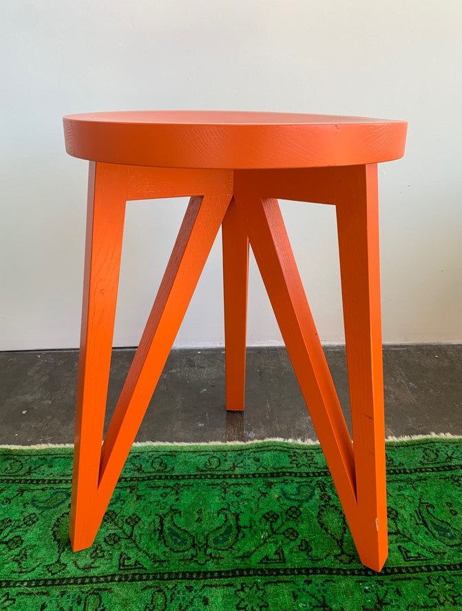 The JL1 Faber Stool by Loehr is a distinctive bright orange wooden piece from the FABER collection. Its round seat and three triangular legs sit on a green patterned rug, showcasing northern European style against a plain white wall.