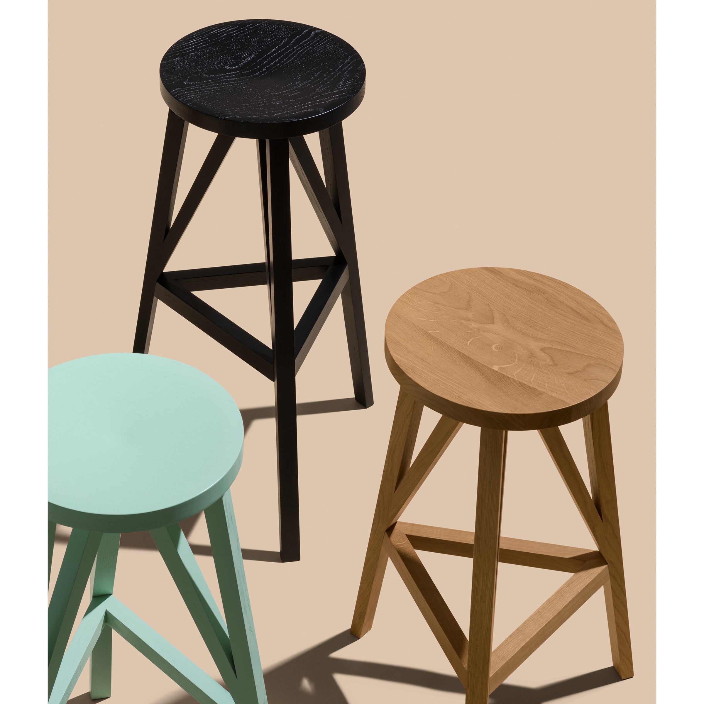 On a beige background, three JL4 Faber Barstools by Loehr stand with round seats and tripod legs, offering a unique silhouette. Black, natural wood, and mint green colors create geometric and color contrasts inspired by northern European architecture.