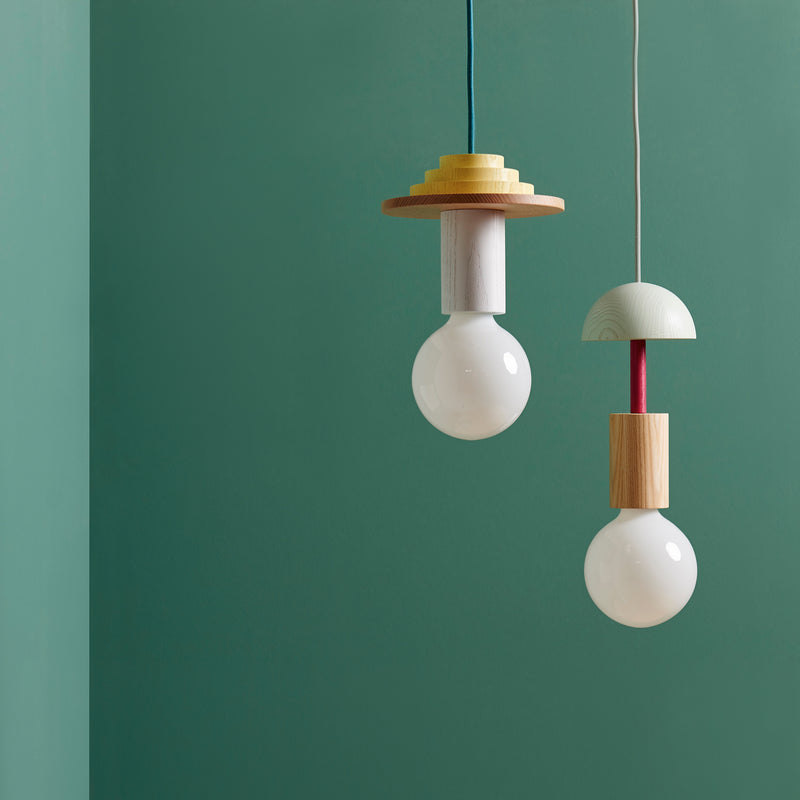 Two modern pendant lights hang against a teal wall. The left boasts layered circular accents, while the right is a Junit Drop by Schneid, featuring ash wood and spherical elements for a minimalist, customizable design in any space.