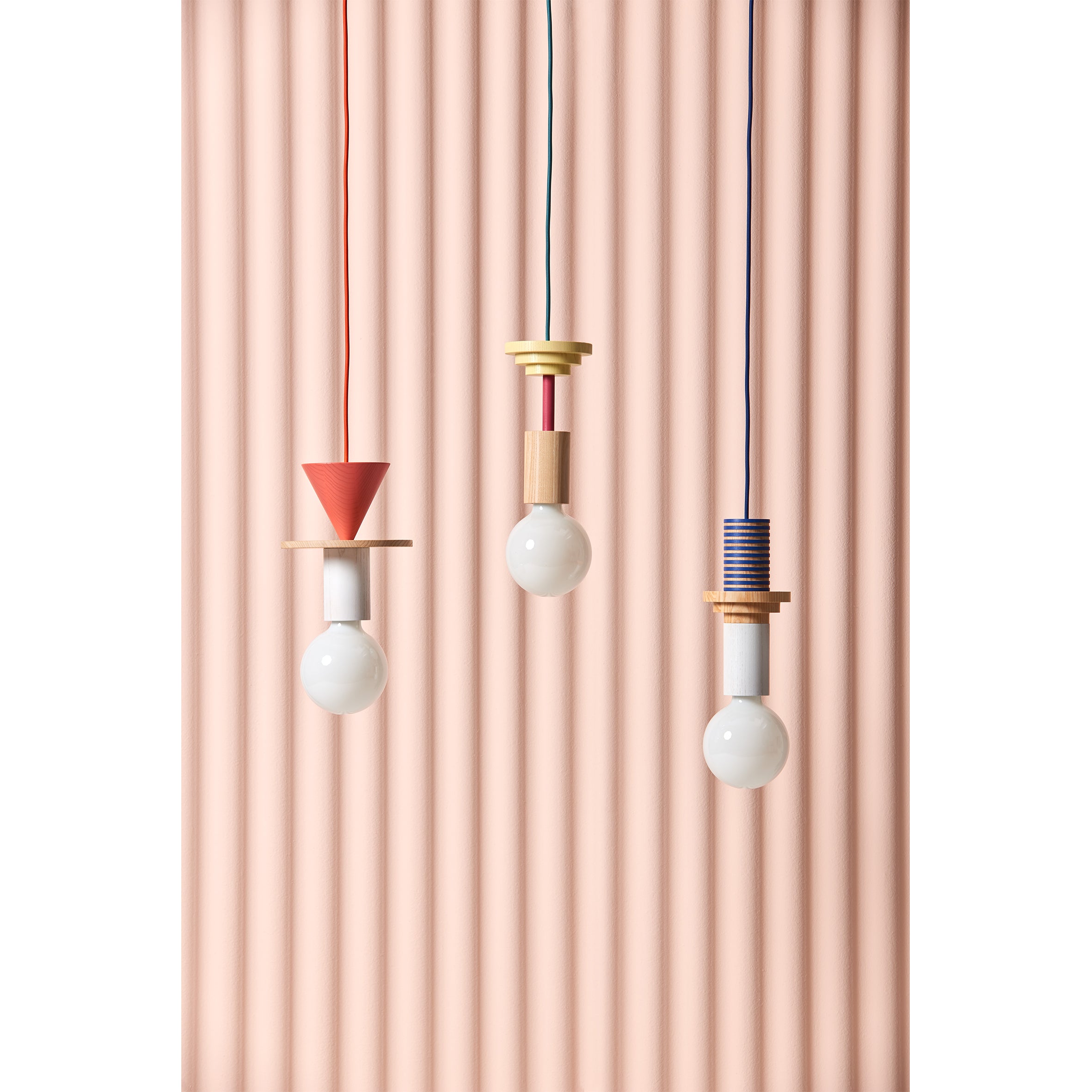 Three Junit Mentis pendant lamps by Schneid, featuring exposed bulbs and geometric designs with accents of a red cone, wooden disk, blue striped cylinder against a pink backdrop, complete with ash wood touches.