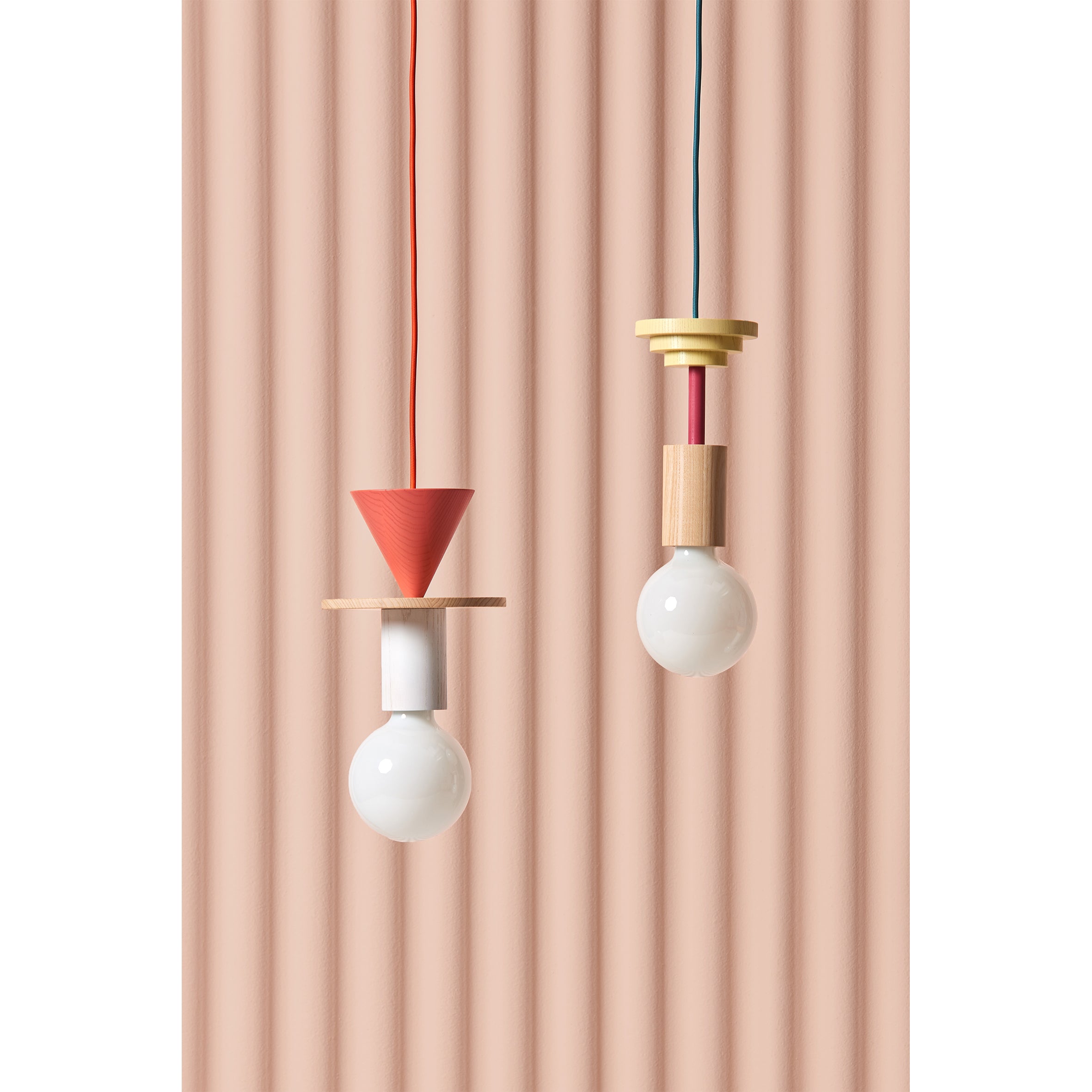 Two modern pendant lights hang against a textured pink background. The left, resembling the Junit Mentis by Schneid, has a red cone and beige disk; the right boasts a modular look with yellow disks and an ash wood cylinder. Both feature exposed white bulbs.