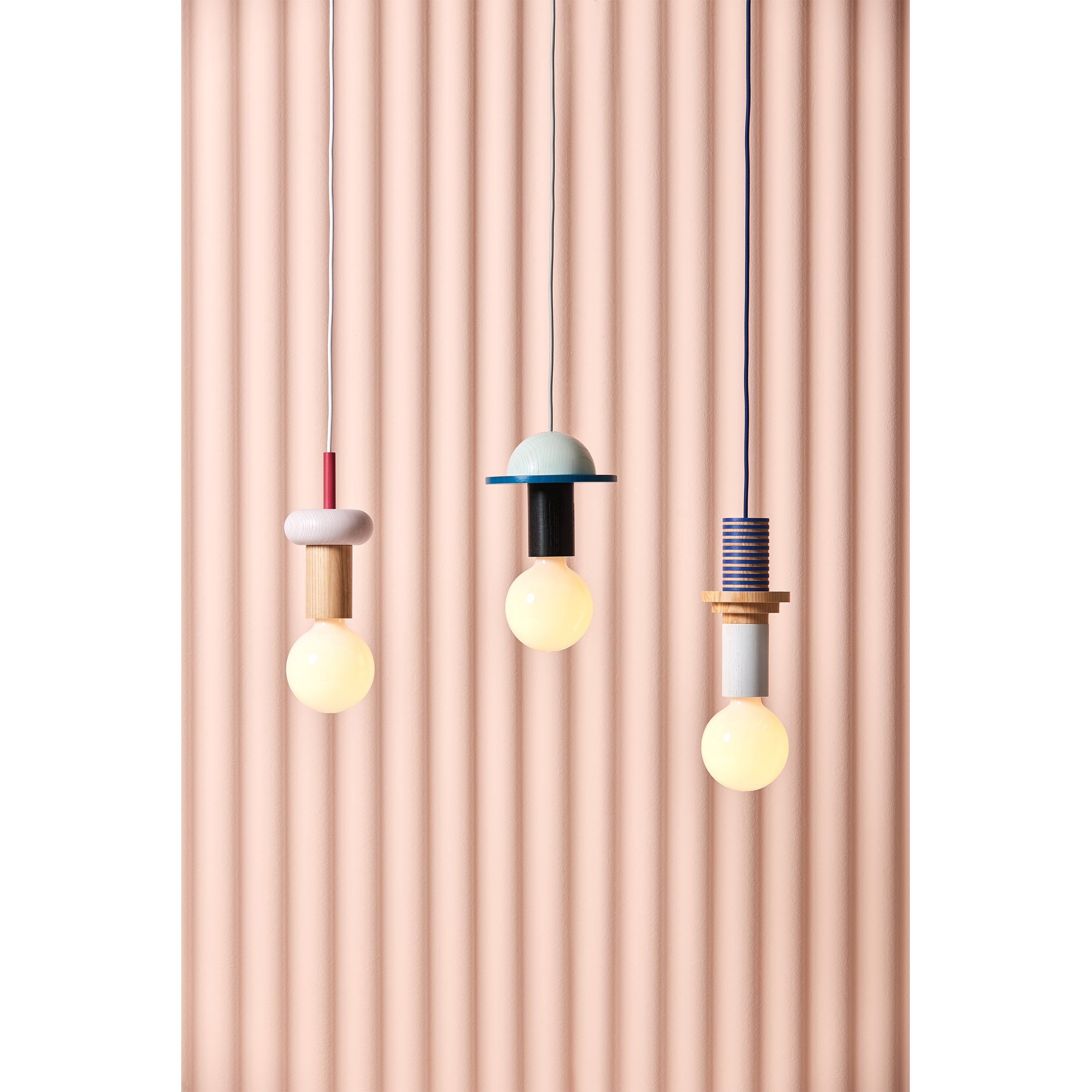 Three Schneid Junit Crescent pendant lamps with colorful geometric designs hang against vertical beige stripes. Each features unique shapes and colors, including red, blue, gold, and silver tones on ash wood encasing warm glowing bulbs.