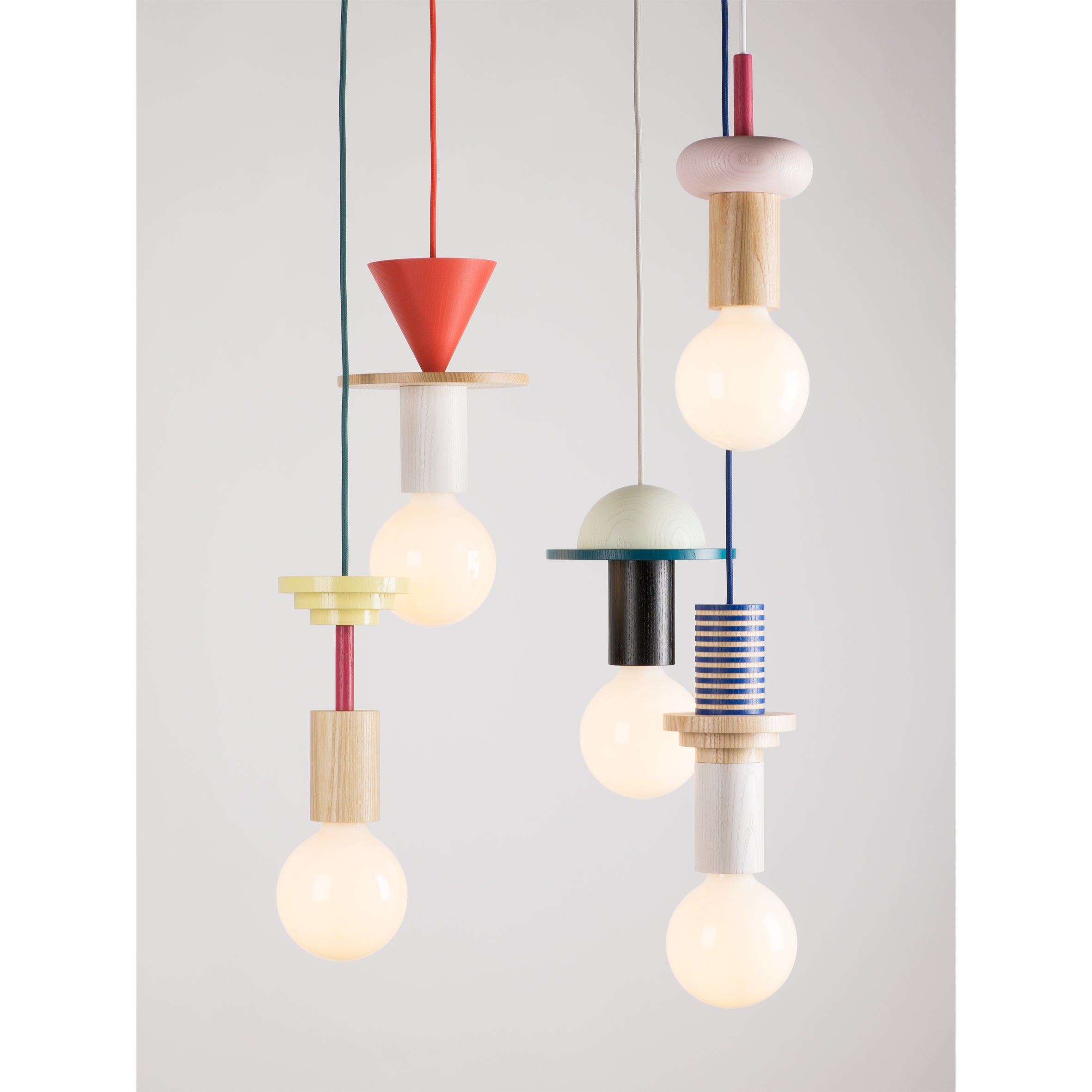 The Junit Mix by Schneid is a set of five modular pendant lamps hung at various heights. Each lamp features unique geometric and colorful designs with ash wood elements, showcasing round bulbs in fixtures like cones, cylinders, and rings for customizable lighting.