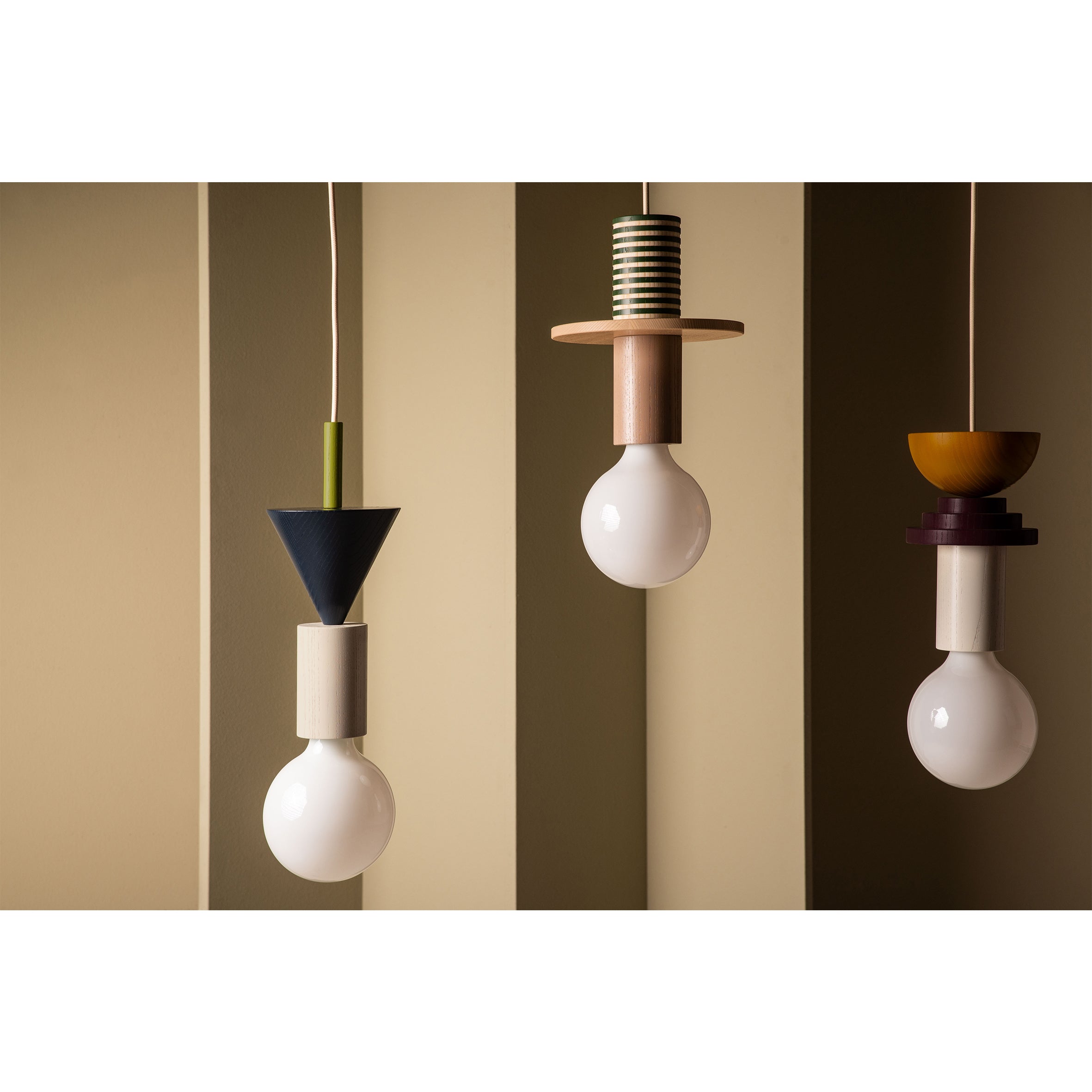 Three Junit Karma pendant lamps by Schneid, showcasing geometric designs, hang from the ceiling against a beige wall. Each features unique ash wood shapes and colors—cones, discs, cylinders—elegantly holding round white bulbs.
