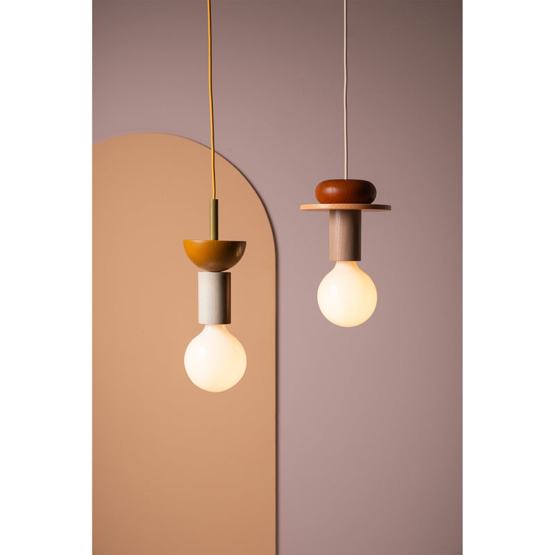 Two Junit Dia pendant lights by Schneid, featuring modern geometric designs in neutral tones, hang against a pink and beige abstract background. Each includes a round bulb and resembles a modular ash wood pendant lamp for added elegance.