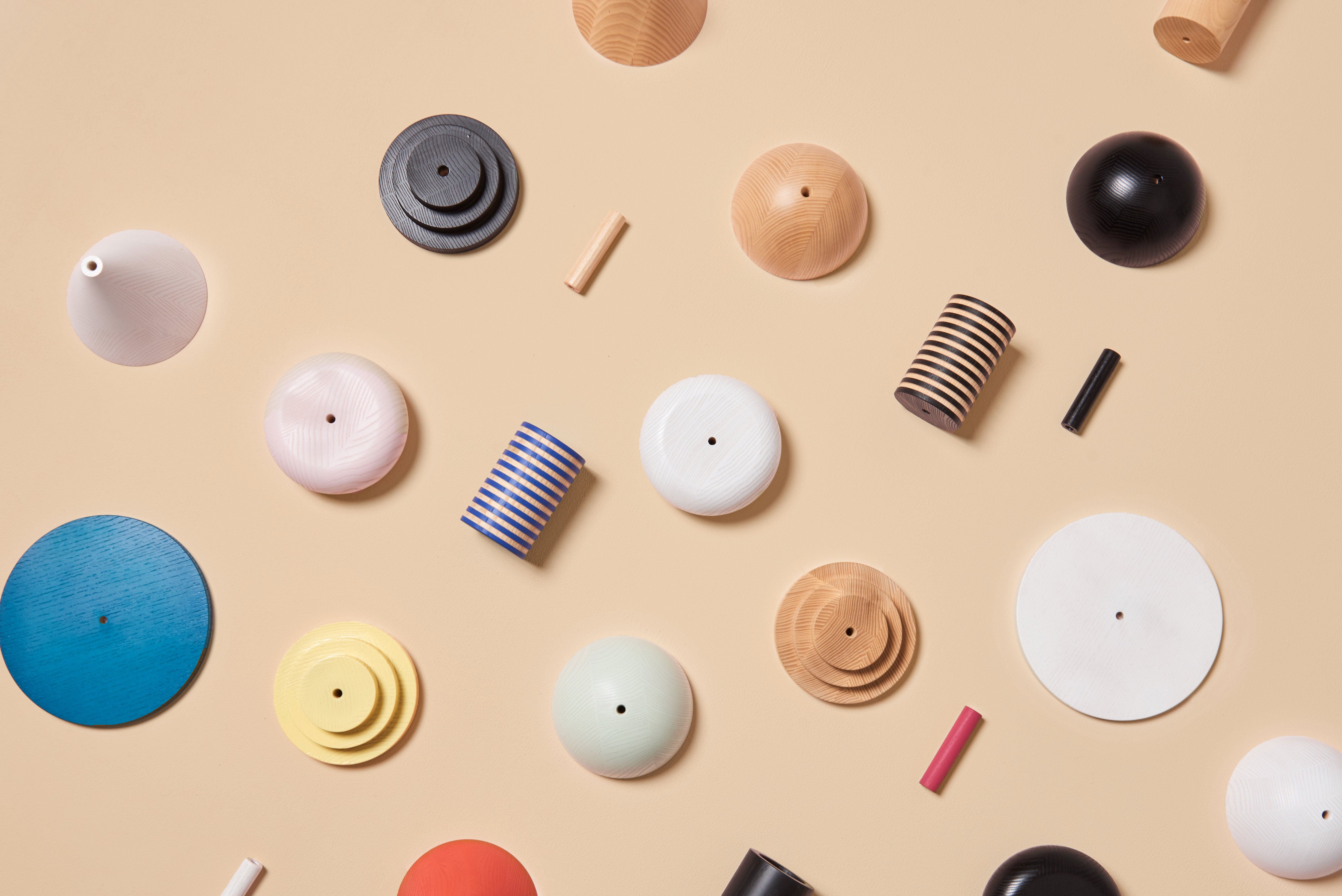 Varied cylindrical and circular ash wood objects, scattered on a beige background, display sizes and colors including blue, black, white, and natural tones. These pieces reflect the charm of a modular pendant lamp and evoke the Junit Column by Schneid.