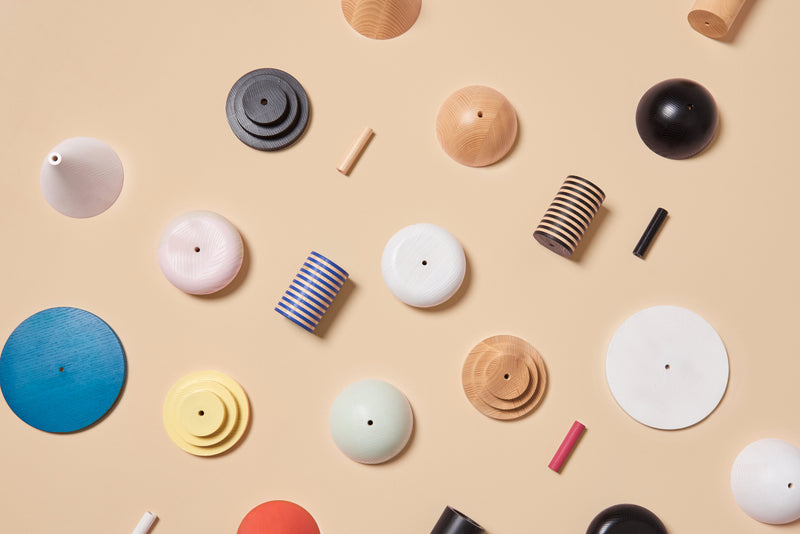 Varied cylindrical and circular ash wood objects, scattered on a beige background, display sizes and colors including blue, black, white, and natural tones. These pieces reflect the charm of a modular pendant lamp and evoke the Junit Column by Schneid.