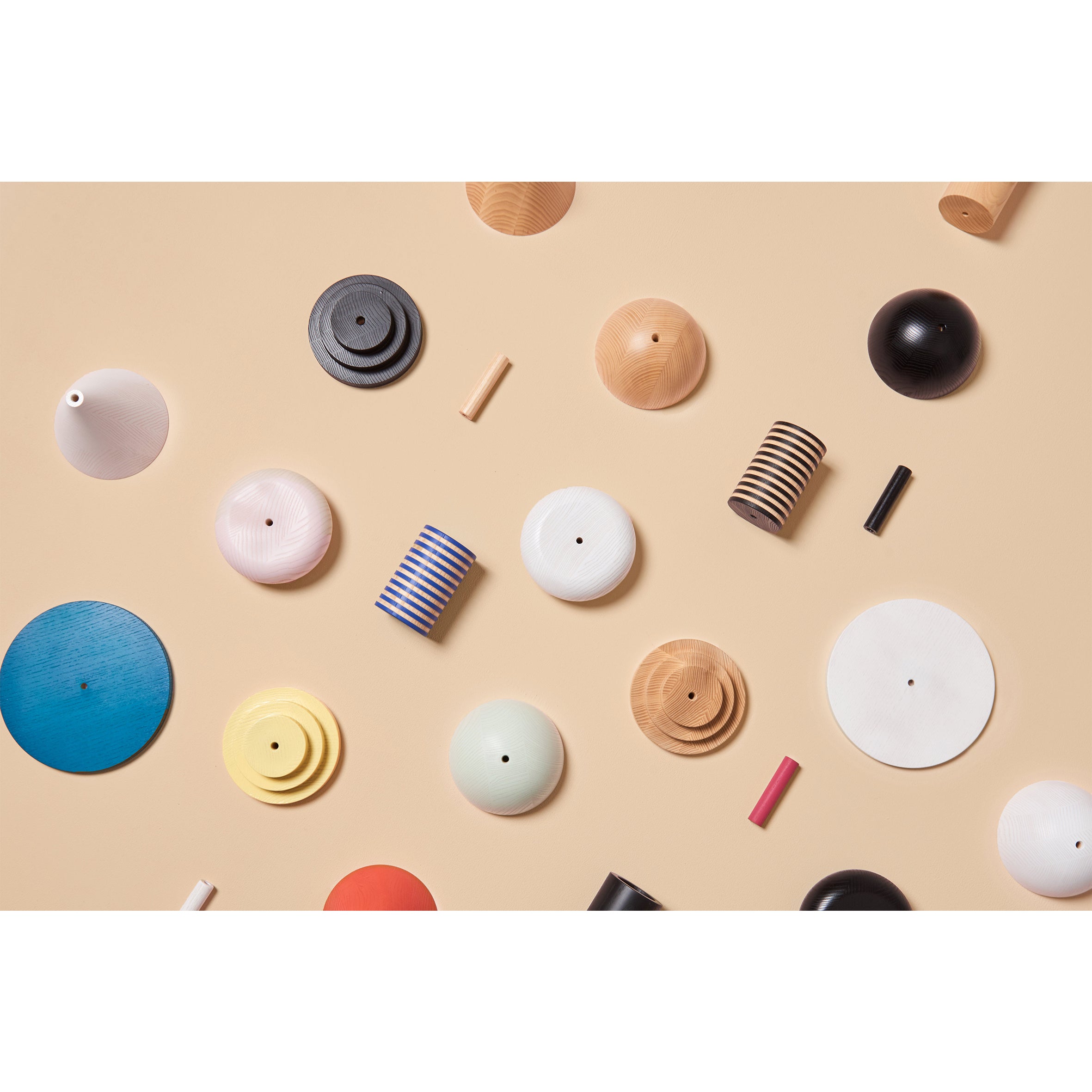 The beige background is scattered with cylindrical and disc-shaped objects from Schneids Junit Drop collection. These pieces, in assorted colors and materials like ash wood and plastic, feature striped and solid patterns that evoke the idea of a modular pendant lamp design.