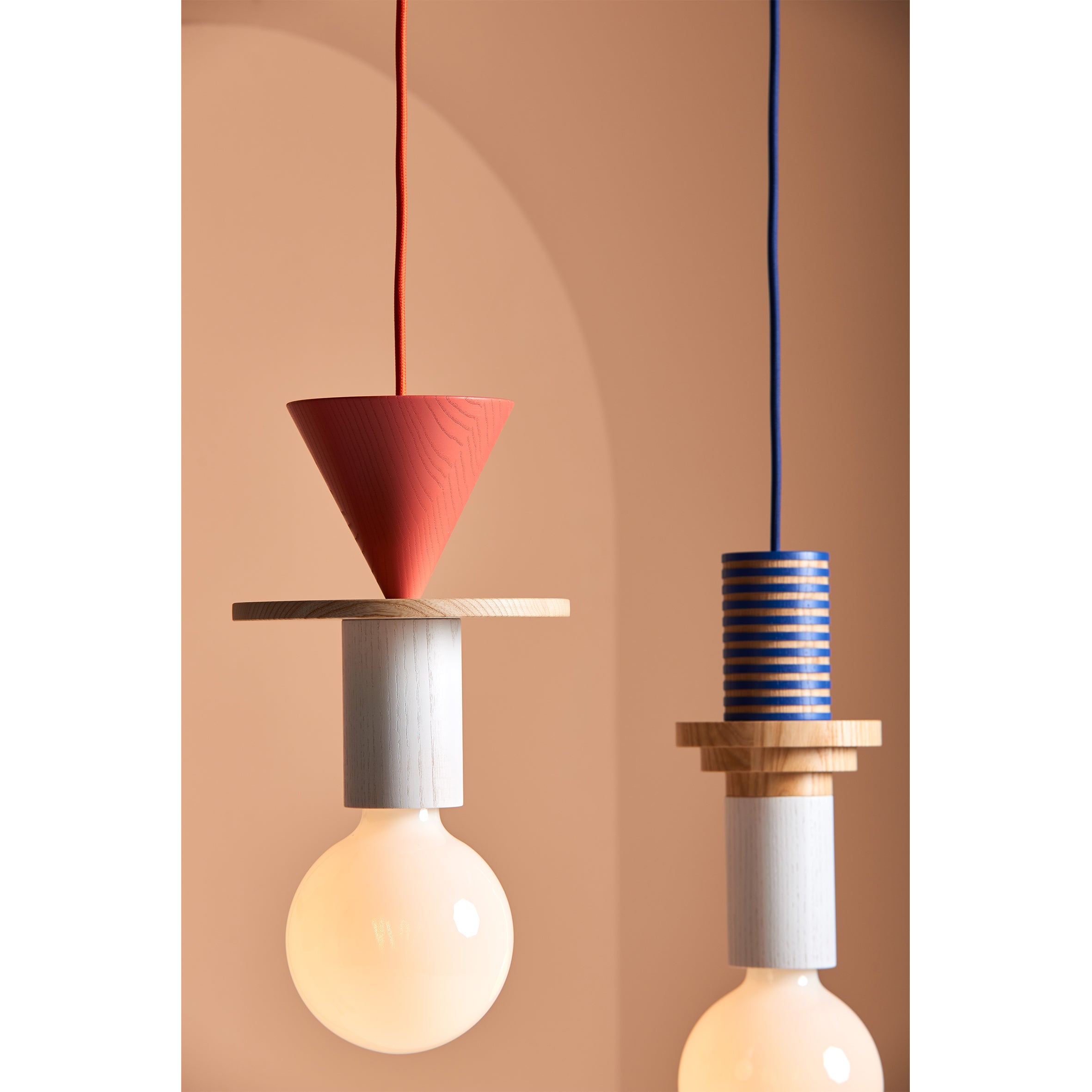 Two modular pendant lights by Schneid feature minimalist geometric designs against a peach background. The Junit Column has a red cone and ash wood disc, while the other sports a blue striped cylinder with a wooden disc. Both have large round bulbs.