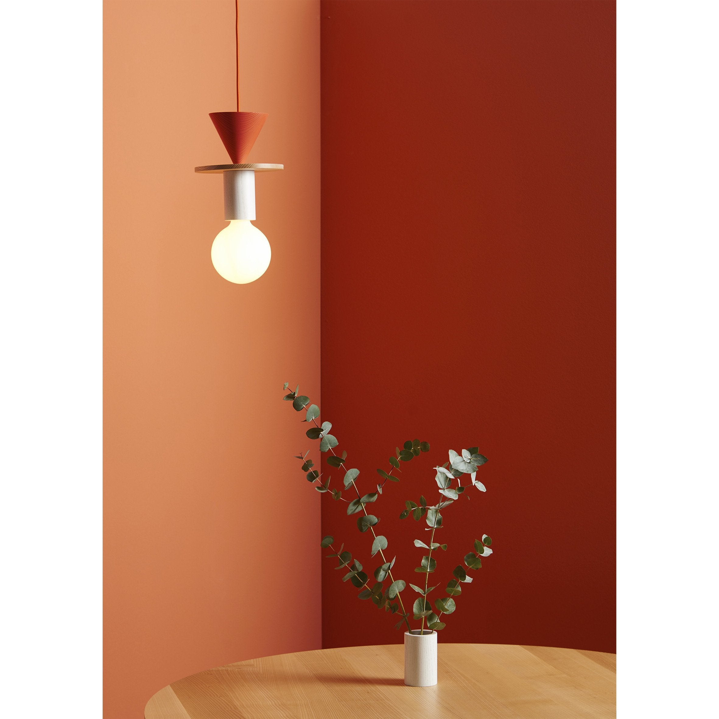 A room with orange walls showcases a Junit Mix modular pendant lamp by Schneid above a light ash wood table. A vase with eucalyptus branches on the table enhances the minimalist, cozy atmosphere, offering customizable lighting to suit any mood.