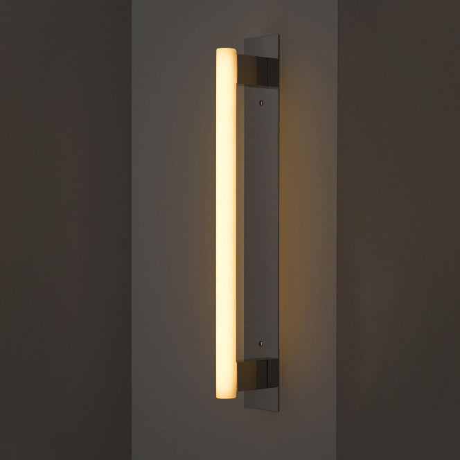The MEA Plate - Sale by KAIA is a sleek vertical wall sconce with a cylindrical glowing light on a brushed brass rectangular base, perfect for dark walls and casting a warm ambient glow.