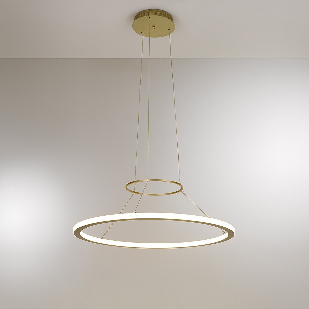 The RIO In and Out Suspension by KAIA is a minimalist pendant light with two suspended rings: an illuminated LED and a decorative solid brass. Thin cables enhance its modern elegance against plain backgrounds.